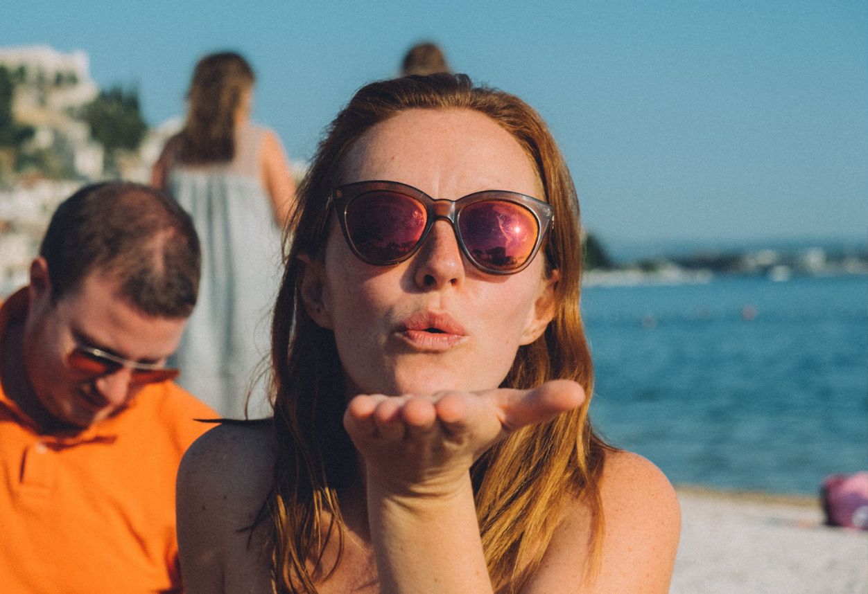 A girl at the beach wearing sunglasses blowing a kiss to the camera