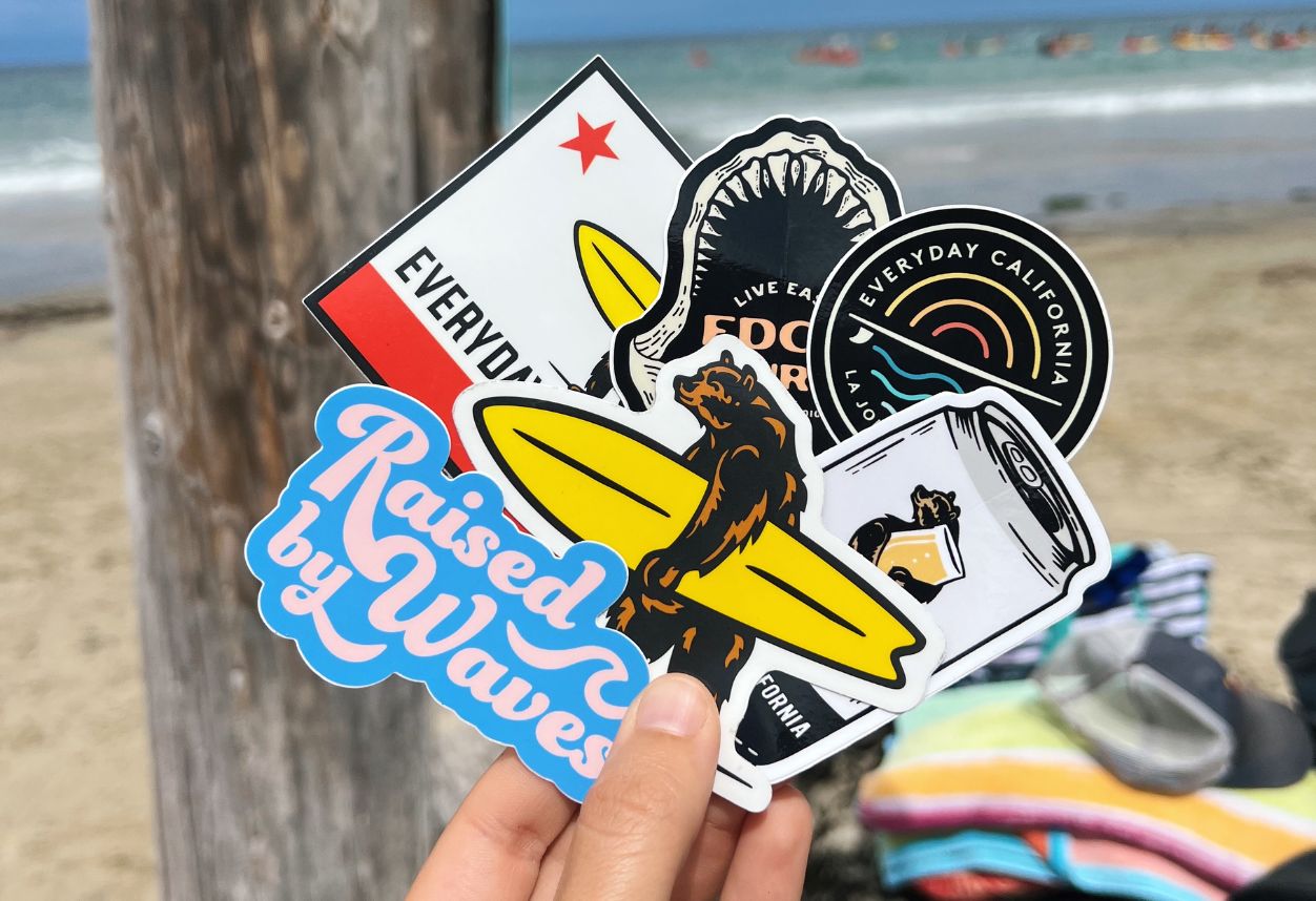 a large pile of everyday California stickers in front of the beach. A bear holding a surfboard, raised by waves, a jaws sticker.