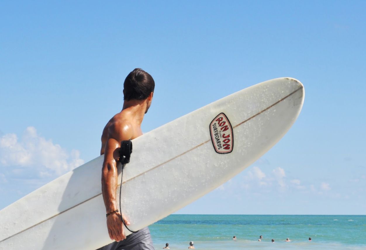 A shirtless surfer holding a white board looking out into the blue ocean