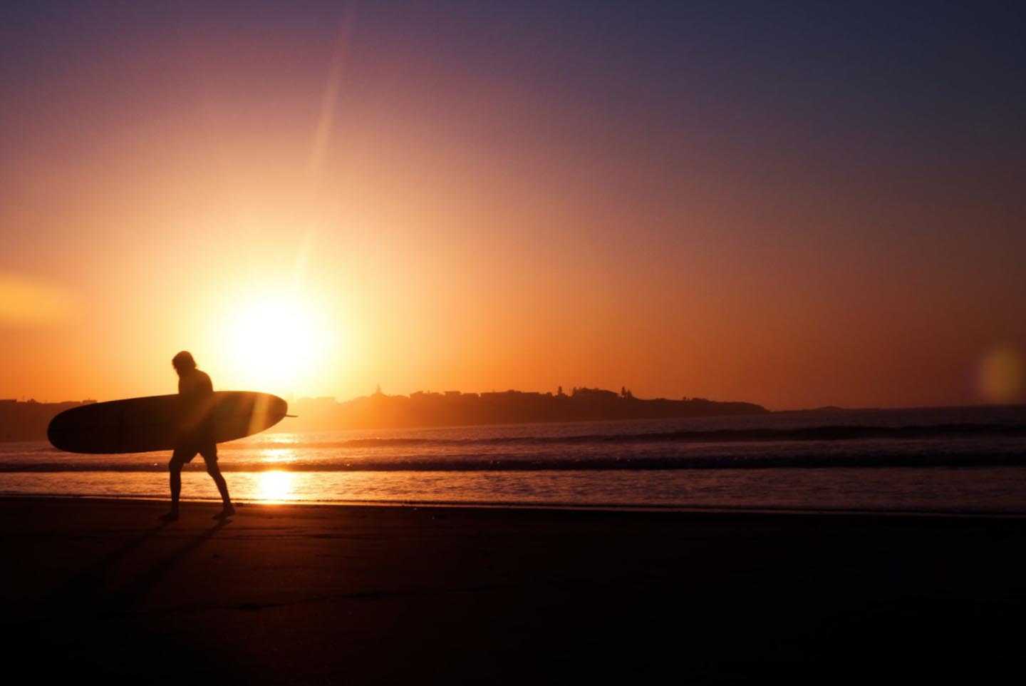 Silueta oscura de un surfista caminando por la playa al atardecer.