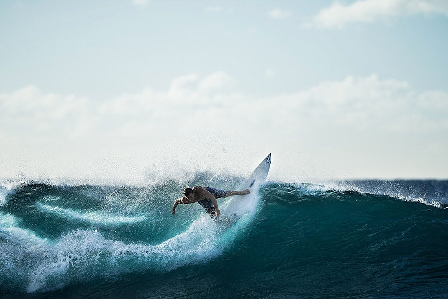 Surfista montando una pequeña ola en el océano. La tabla de surf es blanca y hay nubes en el cielo al fondo.