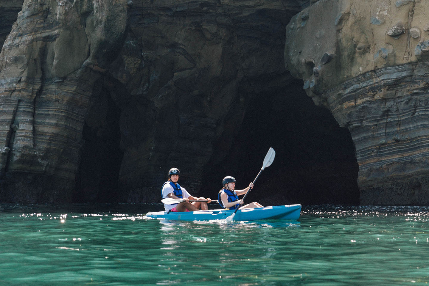 Dos kayakistas en el mismo kayak remando en el océano junto a una cueva marina en La Jolla en un recorrido en kayak por cuevas marinas de Everyday California.