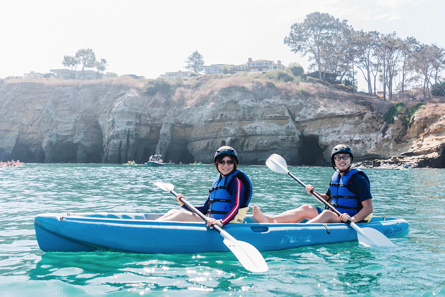 Two kayakers sitting in the same kayak on a sea cave kayak tour in La Jolla with Everyday California. There are no waves in the ocean and there are sea caves in the background of the photo.