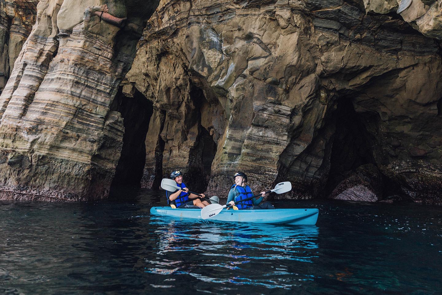 Two kayakers in the same kayak on a sea cave kayak tour with Everyday California, paddling alongside towering sea cave cliffs. The ocean is calm, there are no waves.