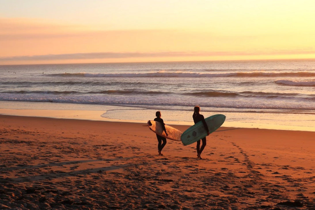 Two surfers holding surfboards on the beach in La Jolla, California. The sun is setting in the background and they are the only people on the beach. The sky is orange and yellow.