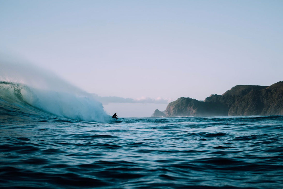 Zoomed out photo of a surfer in southern California, riding a wave. There are sea cliffs on the horizon on the righthand side of the photo. The surfer is the only person in the water.