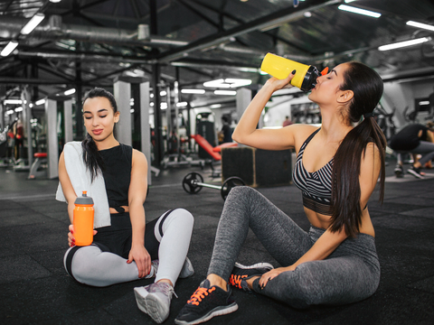2 women at the gym drinking pre-workout dietary supplements to help meet their goals and improve exercise performance.