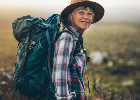 older white woman with no makeup and a friendly expression, wearing a hat, checked shirt and backpack, turning back towards the camera as she hikes.