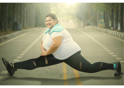 A light skinned woman of East Asian ethnicity with a larger body is doing the splits in the middle of a race track. She is wearing running trainers, black leggings and a white top, and is smiling at the camera.