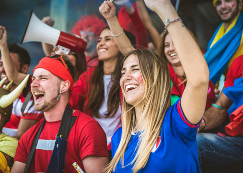 a white woman with long blond hair and a blue t-shirt smiling and raising her hand in a sports stand. Other people dressed in red are cheering around her.