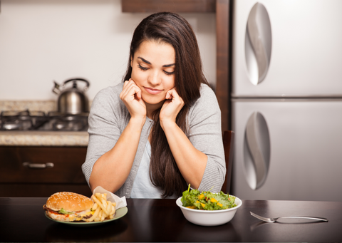 White woman sitting at a table looking at a plate of fast food next to a plate of salad.