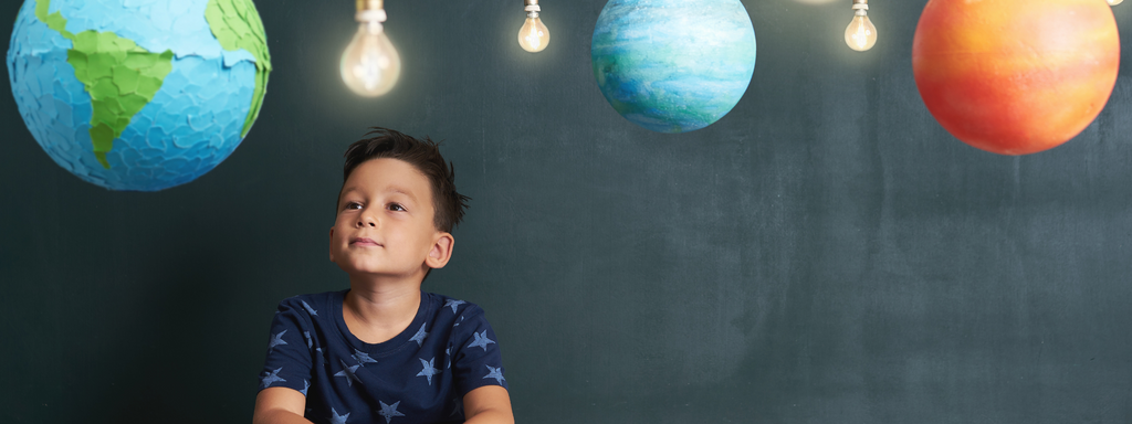 Little boy sitting at a table and looking up, with models of the planets overhead