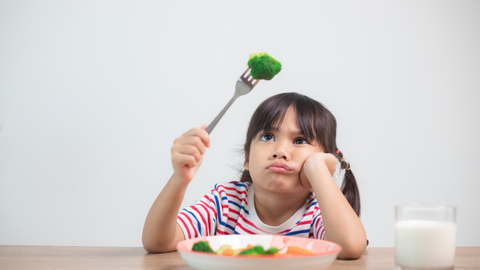 Little girl sitting at a table with a plate of food in front of her and a piece of broccoli on her fork