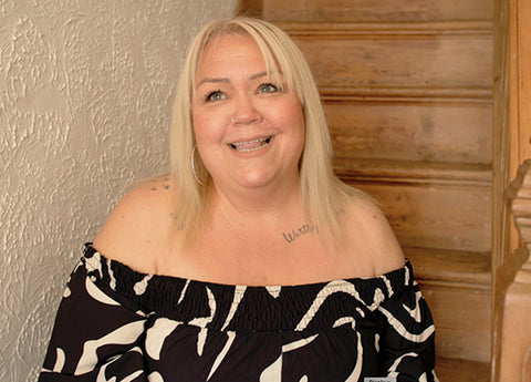 Upper body of a blond, white woman in her 30's smiling happily, wearing an off-the-shoulder black and white top. Her hair and makeup has been nicely done.