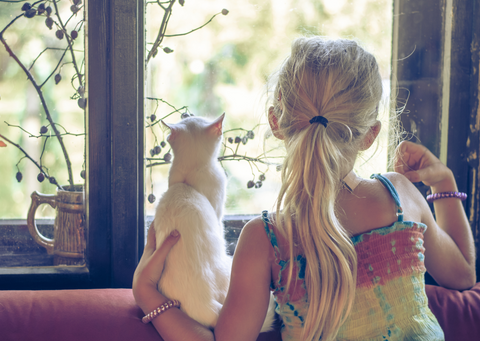 Young white girl with blond hair is seen from behind, looking out of a window. Her arm is around a small white cat who is also looking out of the window.