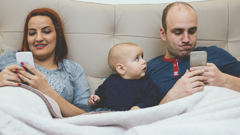 baby in bed between two adults staring at their phones