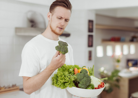 A young white man standing in a kitchen holds a big bowl of vegetables in one hand and a floret of broccoli in the other. He is looking with some confusion at the broccoli.