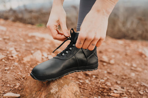 Woman tying her trekking barefoot ankle boots, leaning on a rock