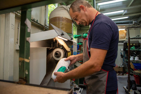 Unser Kollege steht an einer Maschine, hält einen Schuh in der Hand und schleift die Kante, an der die Sohle sitzen wird.