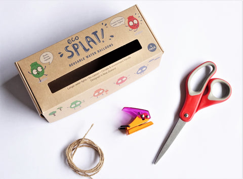 An empty EcoSplat reusable water balloon box on a white background, scissors, string and a holepunch