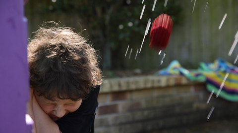 A 9 year old boy hiding behind playground equipment as a red EcoSplat reusable water balloon is thrown towards him. Water is splashing around.