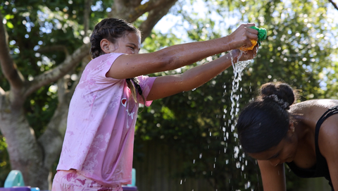 A 12 year old girl squeezing a EcoSplat reusable water balloon onto her friend's head. Water is falling onto her friend's head. They are laughing and having fun.