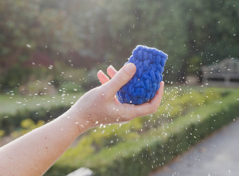 A hand holding a blue EcoSplat reusable water balloon in front of a garden. There is water splashing and light is shining on the droplets