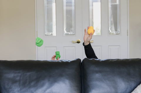 Two hand popping out from behind a black sofa and throwing dry EcoSplat reusable water balloons.