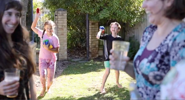 Two women standing in a garden holding drinks and laughing. A boy and a girl are in the background ready to throw a blue EcoSplat reusable water balloon towards themEco friendly water fights for your eco home this summer. 