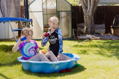 Two 10 year old girls sitting in a paddling pool in a sunny garden. They are holding EcoSplat reusable water balloons ready to throw them and are laughing. Lying on the grass are red EcoSplat reusable water balloons