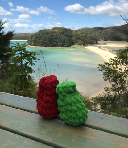 A red EcoSplat Reusable water balloon and a green EcoSplat reusable water balloon upright on a bench overlooking a sunny beach with forested hills around. The reusable water balloons are leaning on each other reminiscent of a couple leaning against each other romantically.  