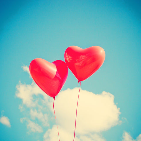 Two red balloons in the shape of hearts on strings against a blue sky with clouds in it.