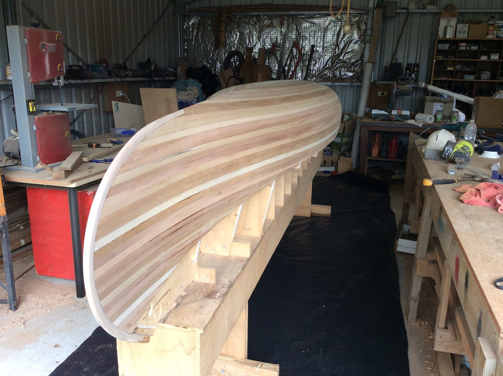 A sanded wooden canoe hull, unfibreglassed and sitting on the strongback