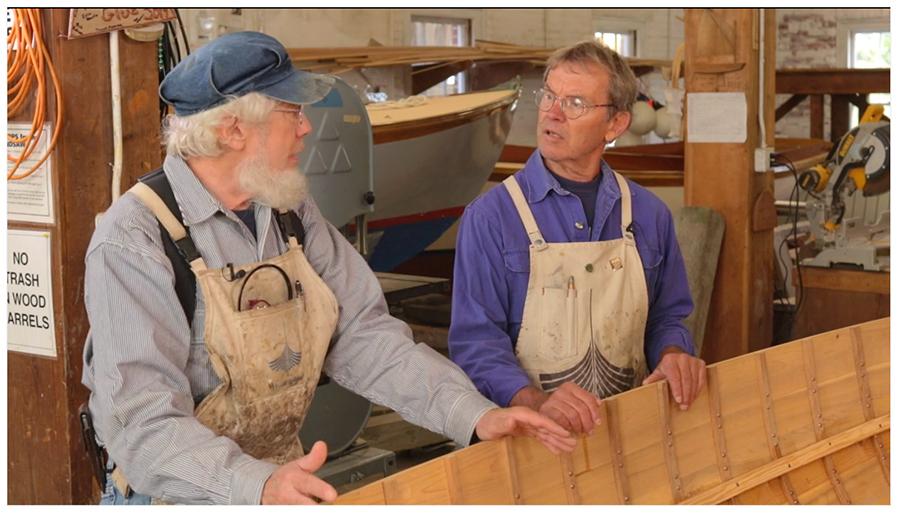 Two boat builders in carpenter's aprons stand with their hands on an unfinished hull