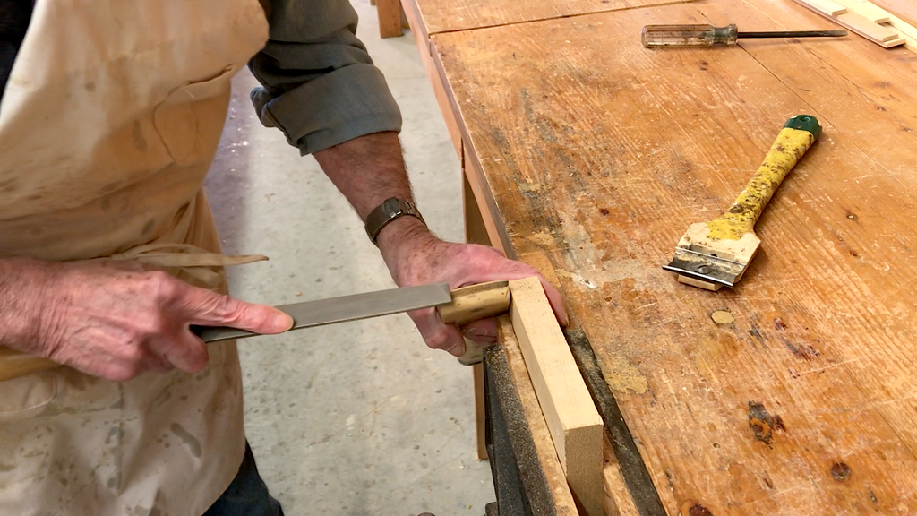 Ted Moores files the blade of a Richard paint scraper to a custom curvature, with the scraper braced against a work table and a second scraper seen beside it