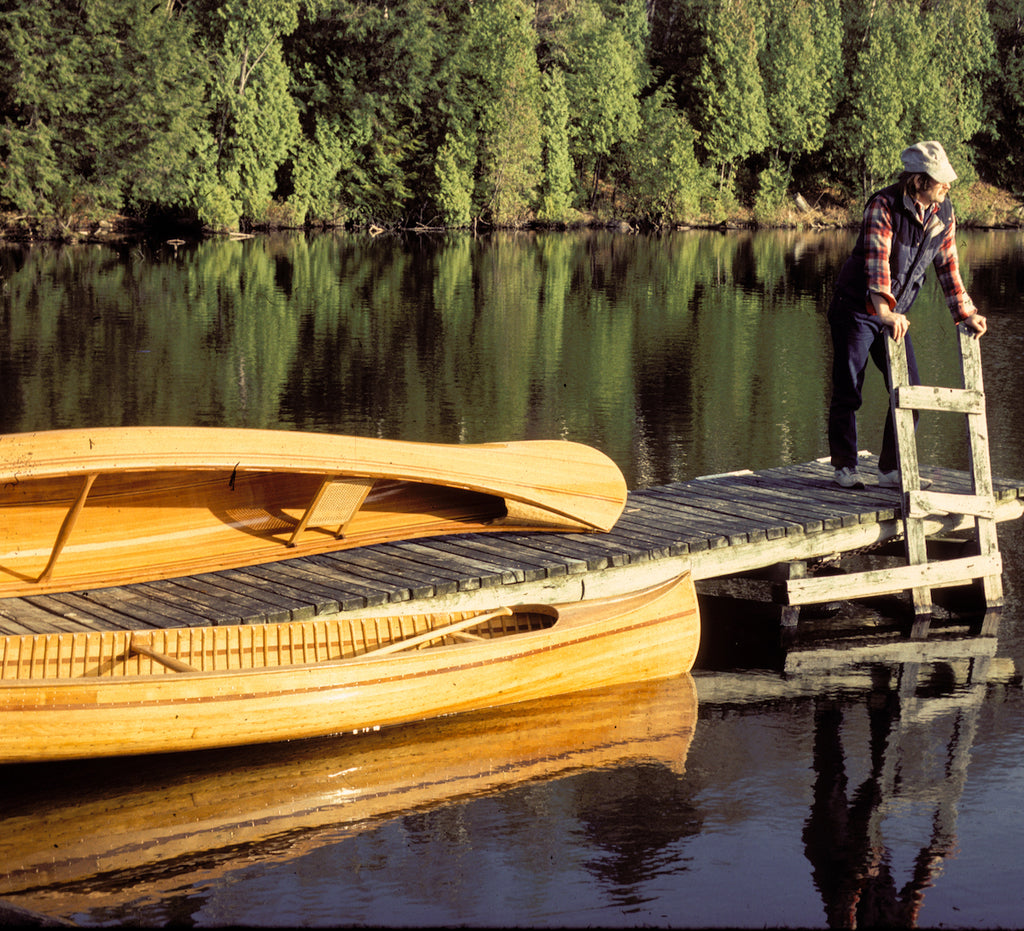 Ted Moores leans on the ladder of a dock, with two examples of woodstrip canoes beside him (recognizable as similar to the cover of Canoecraft to fans of the book)
