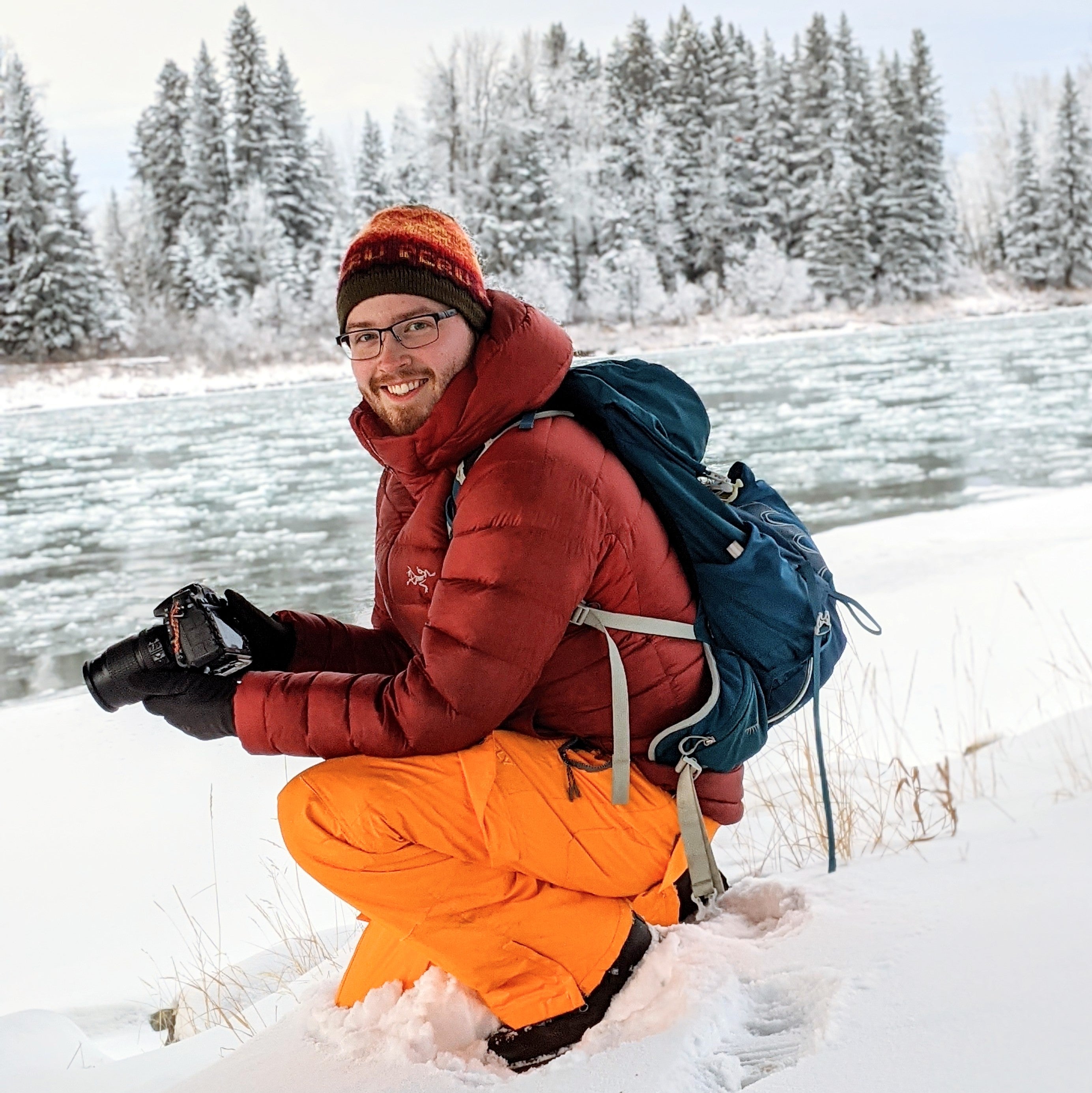 Geoffrey Coulthard Photographer About Me. Portrait or Profile picture of Geoffrey coulthard. Photographer for GeoffreyPrints. Kneeling in snow with orange snowpants and red jacket holding a camera. Kneeling in front of a flowing river covered with ice chunks and snowy trees in background. 