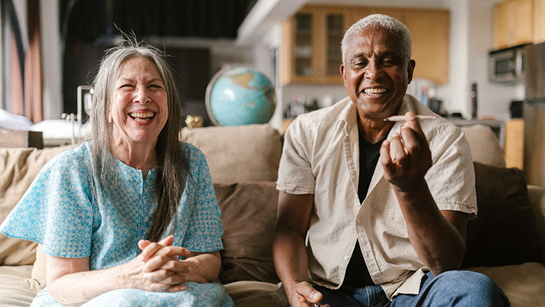 Multiethnisches, älteres Paar sitzt auf einem modernen braunen Sofa und raucht lachend einen Joint.