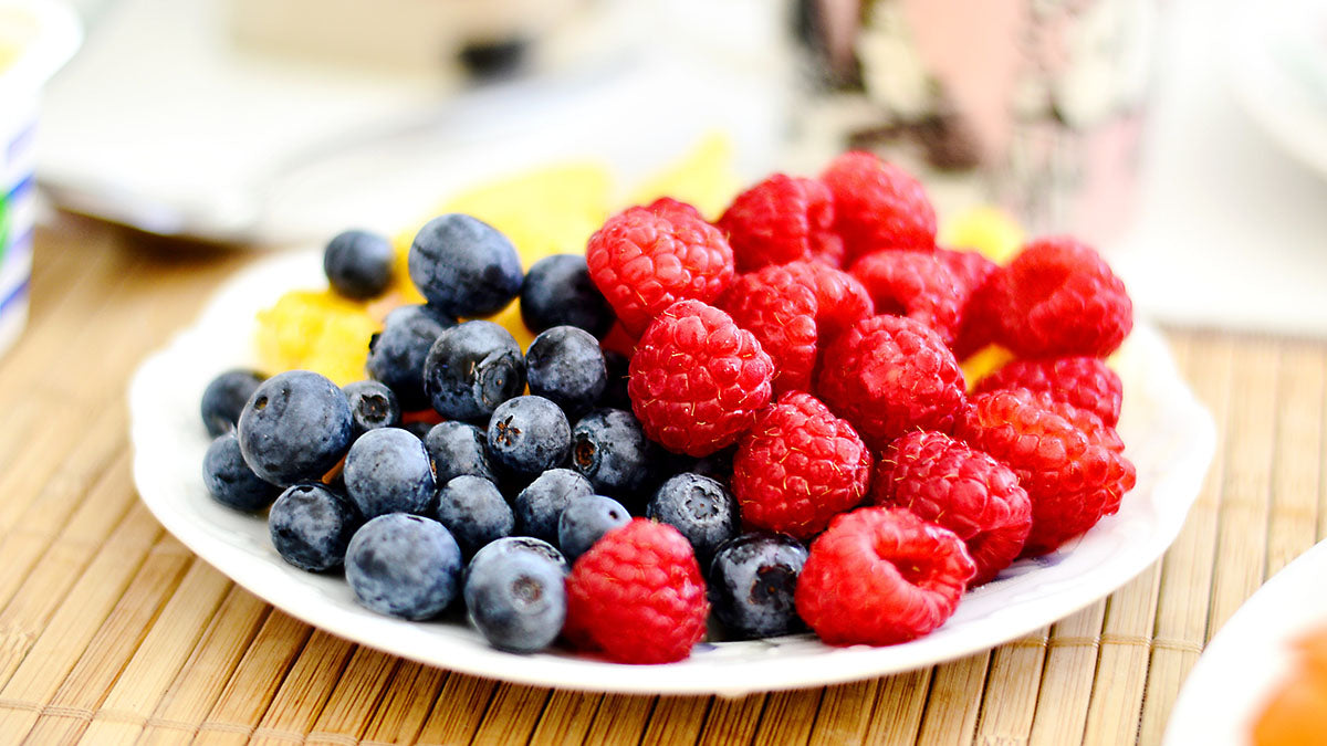 Ein Beeren Mix aus Heidelbeeren und Himbeeren ist auf einem weißen Obstteller aus Keramik platziert, der auf einer hellbraunen Bambusmatte steht.