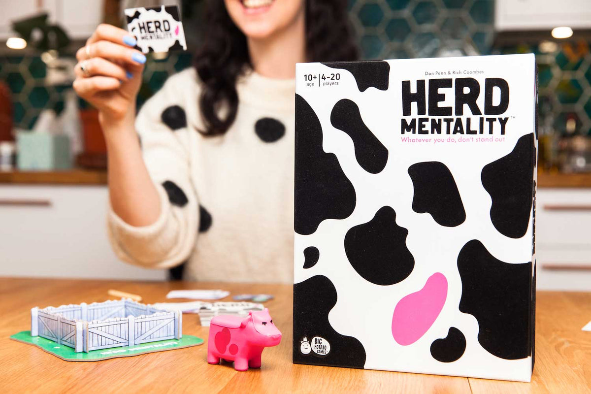 Herd Mentality game box, pink cow, paddock, and person reading a question in the background.