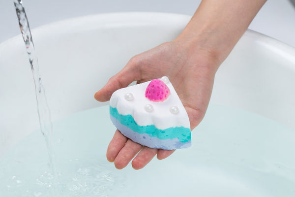 a hand holding a cake shaped bath bomb over a bathtub 