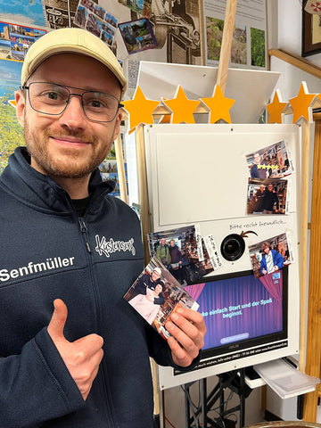 Andy steht neben der Fotobox. Er hält ein Bild in der Hand und zeigt einen Daumen hoch. Er trägt eine blaue Jacke und eine beige Schirmmütze.