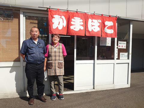 赤地に白で「かまぼこ」と書かれたのれんの下に立つ青い服の男性と茶色いチェックのエプロンをした女性の写真