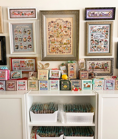 The Frosted Pumpkin Stitchery's Office. Framed cross stitch pieces are on the wall, cross stitch cubes are on the top of the shelves and bins are filled with kits.