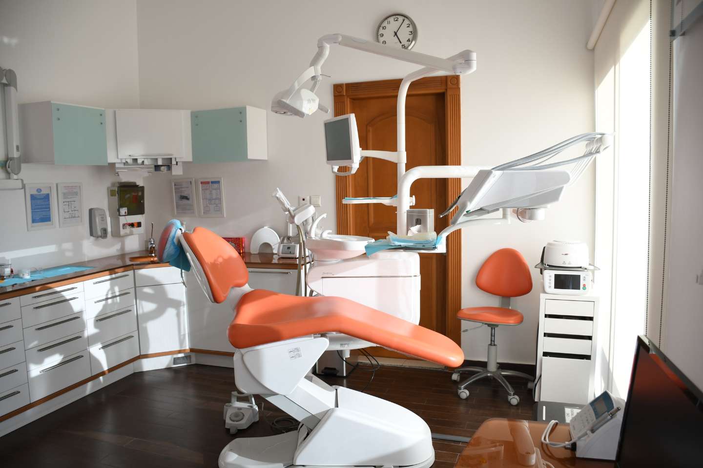 A typical dental surgery, white cabinets line the back and side walls, with a large orange bed-chair in the middle of the room. Small orange chair in the corner with large dental equipment above the chair.