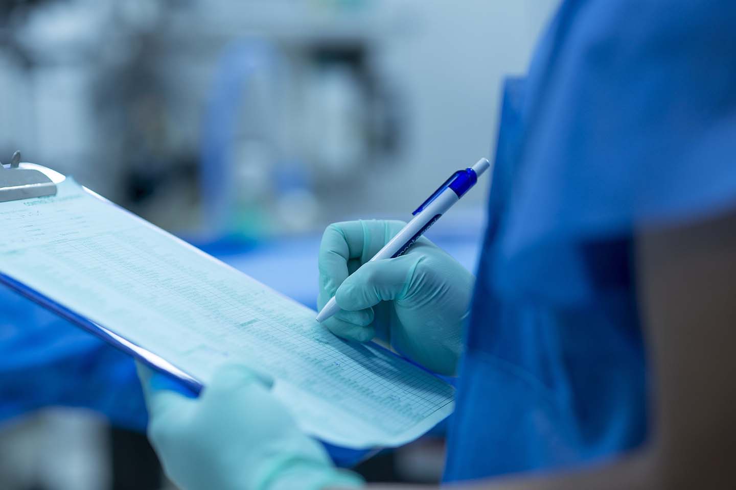 A medical professional wearing scrubs and blue gloves while writing on a patient's chart.