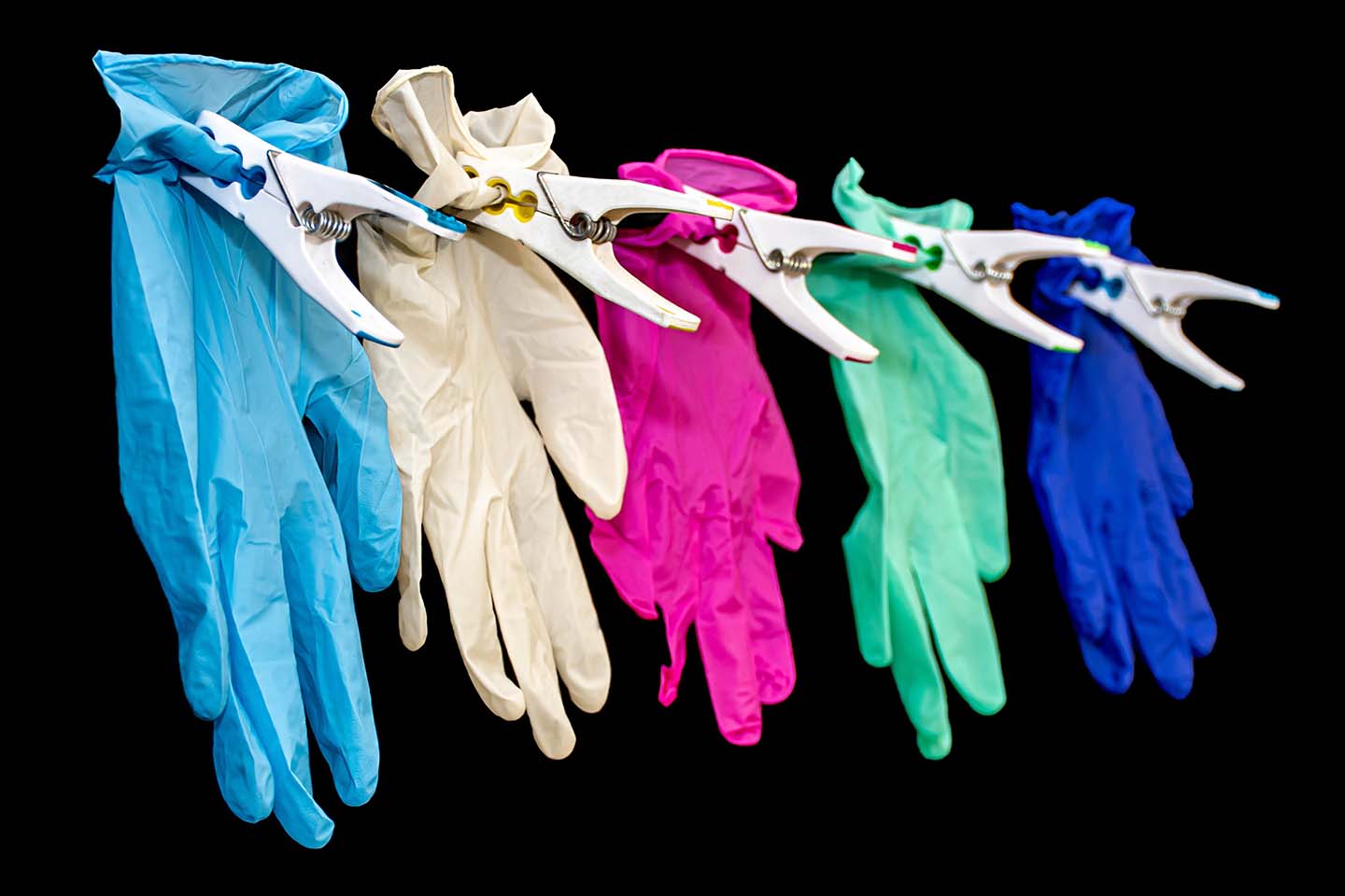 Five differently colored surgical gloves pinned to a wall against a black background: sky blue, white, pink, green, and blue.