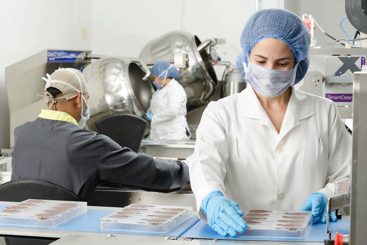 Woman in a lab with other people in the background wears PPE including blue medical gloves.