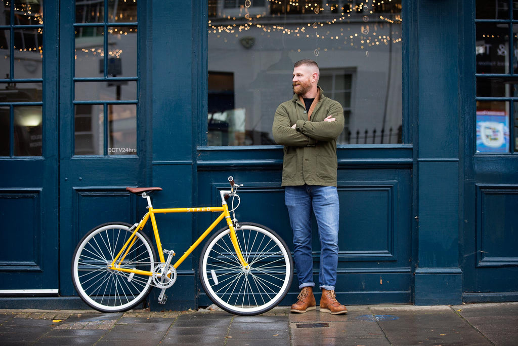 man standing next to yellow single speed bike against dark blue wall