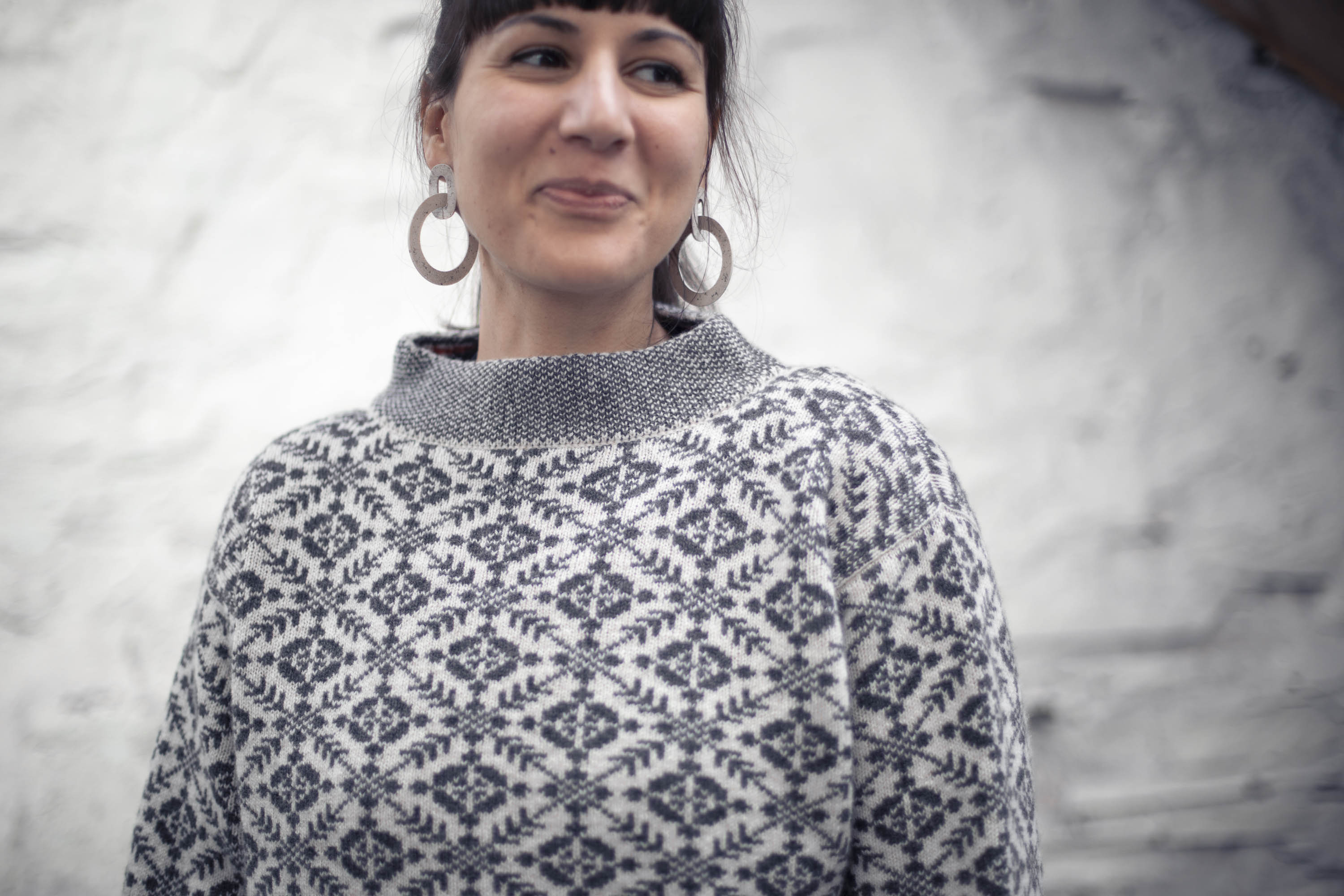 A woman with olive skin and dark hair tied back wears a charcoal grey and white fair isle style jumper with a stand up neck. She smiles and looks to one side.